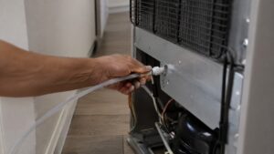 A hand connecting a thin water tube to the back of a refrigerator pulled out from a kitchen wall