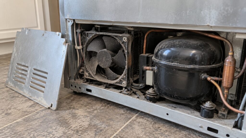 A small black fan with plastic blades sits behind a removed panel near the compressor at the back of a refrigerator, coated in grey dust